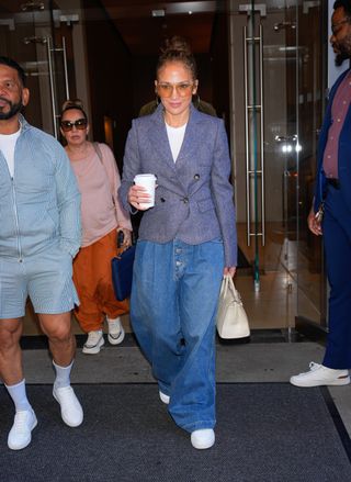 Jennifer Lopez on July 26, 2024 in New York City wearing a blazer, a white t-shirt, pleated jeans, white sneakers, aviator sunglasses, and an ivory bag.