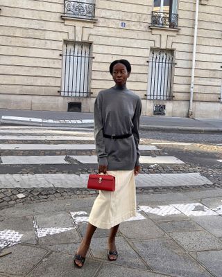 Sylvie Muse wearing a gray sweater, a black belt, a midi skirt, black heels, and a red Savette bag.