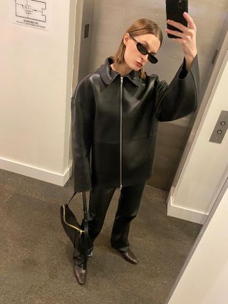 Fashion influencer @christietyler with slicked hair taking a mirror selfie outside an elevator wearing a minimalist black collared leather jacket, black trousers, boots, and sunglasses.