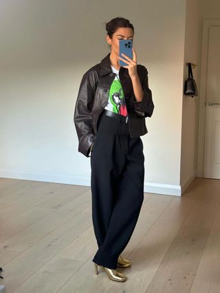 Fashion influencer @smythsisters taking a mirror selfie wearing an on-trend leather jacket, a graphic tee, beltered trousers, and metallic boots.