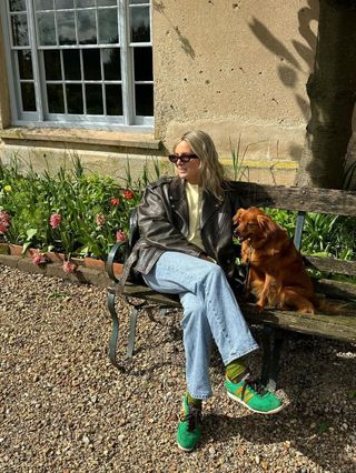 Fashion influencer @lucywilliams02 sitting outside on a park bench with her dog wearing an oversized leather biker jacket, blue jeans, and green New Balance sneakers.