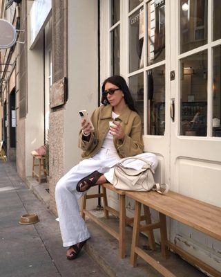 British influencer Jessica Skye poses while looking at her phone on a bench outside a UK cafe wearing a camel canvas coat, white top, black oval sunglasses, white linen pants, a Loewe puzzle bag, and brown suede sandals