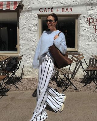 British influencer Jessica Skye poses with a smile in front of a cafe in Cornwall wearing a brown suede tote bag, a light blue sweater, striped linen pants, and sneakers