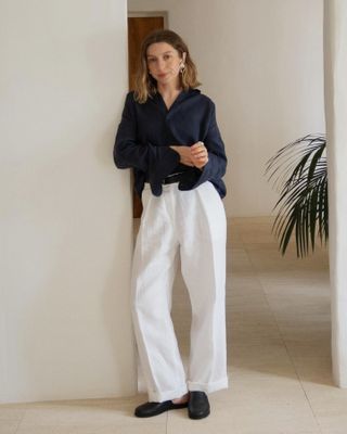 British influencer Brittany Bathgate poses in silver shell earrings, a navy button-down shirt, black belt, and white linen trousers