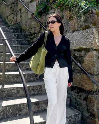 British influencer Jessica Skye poses in black oval sunglasses, a black button-down cardigan, green suede tote bag, and white drawstring linen pants