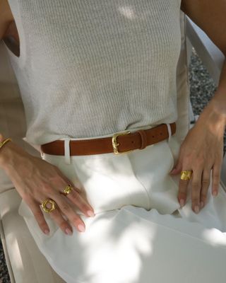 Cass Dimicco wears a white tank top, white pants, tan belt, and gold rings.