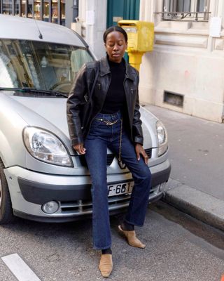 Sylvie Mus wears a black leather jacket, black turtleneck sweater, blue jeans, tan mules, and a gold chain belt.