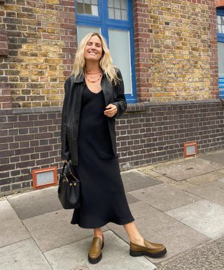 British influencer Lucy Williams smiles while posing on a London sidewalk wearing layered beaded necklaces, a black leather jacket, black slip dress, black bag, and olive green chunky loafers