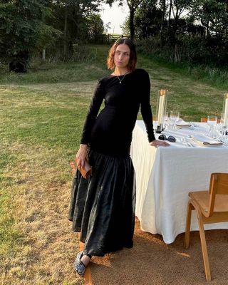 influencer Erika Boldrin poses next to an outdoor dining table in Somerset, England wearing a black long sleeve drop-waist dress and studded Mary Jane flats