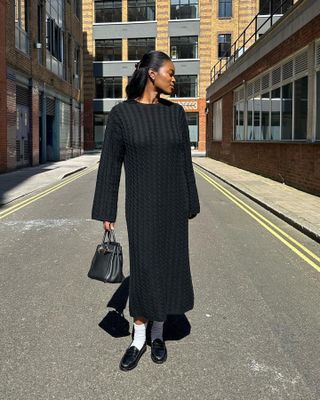 British influencer Marilyn Nwawulor-Kazemaks poses on a street in London wearing a cableknit maxi sweater dress, Hermes bag, white crew socks, and black loafers