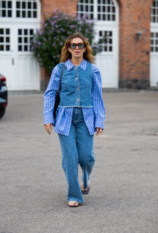 Woman walking after show wearing blue button-down shirt, denim waist-coat, jeans and flip-flops during Copenhagen Fashion Week.