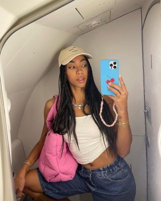 Dutch fashion influencer Amaka Hamelijnck taking a selfie on an airplne wearing a white tank, baseball hat, denim shorts, and pink tote bag