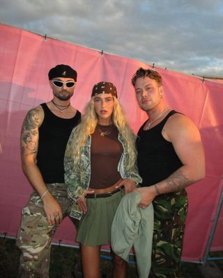 Emili stands with two men wearing a green pleated miniskirt, a black belt, brown top, camo jacket, brown bandana, and gold necklace.