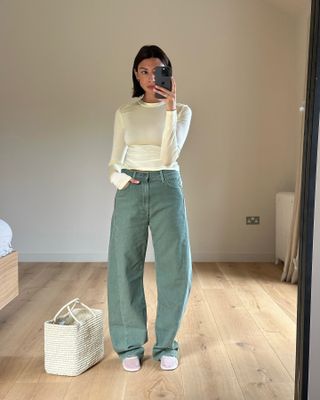 A woman wears green jeans, white mesh flats, a white woven tote bag, and butter yellow long-sleeve top.