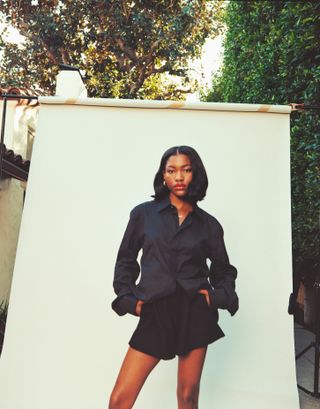 Demi Singleton stands in front of a cream seamless wearing a navy button-down shirt and black shorts.