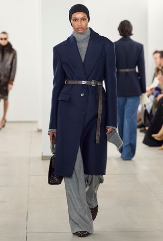 the navy color trend shown on Michael Kors's fall runway worn by a model wearing a navy blue coat over a gray sweater with matching gray pants, a brown belt, and brown pointed heels