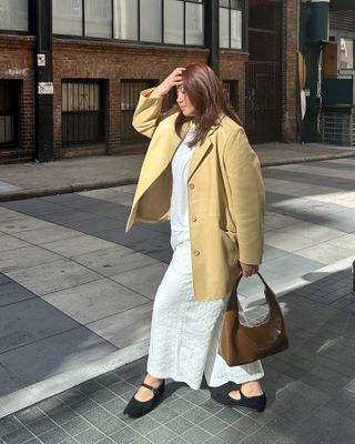 female fashion influencer poses on a city sidewalk wearing a butter yellow leather blazer jacket, white tee, white linen pants, brown shoulder bag, and black Mary Jane flats