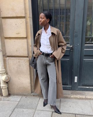 female fashion influencer and stylist Sylvie Mus poses on a Paris sidewalk wearing a brown trench coat, white button-down shirt, a black belt, gray straight-leg jeans, sheer black tights, and black slingback heels