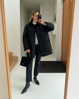 female fashion influencer Anouk Yve poses for a mirror selfie wearing a black quilted coat, cardigan sweater, straight-leg ankle jeans, mini black tote bag, and black pointed-toe boots