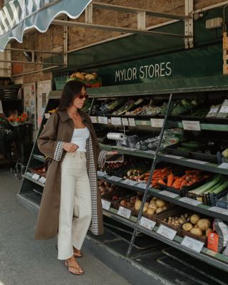 An influencer wears a trench coat, beige jeans, beige flip-flops, a white tank top, and red sunglasses.