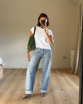 Influencer takes mirror selfie wearing white t-shirt, cuffed jeans, and suede green bag.