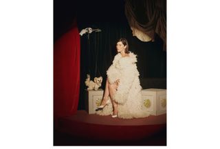 Gracie Abrams wearing a cream-colored furry Stella McCartney long coat with matching Ferragamo heels sitting on a painted chest next to a dog puppet for Who What Wear's fall issue.