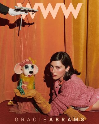 The cover of Who What Wear's fall issue starring Gracie Abrams in a pink, orange, and yellow F/W 24 Miu Miu look. She is posing with a puppet in a similar color-blocked outfit. On the cover, it says "WWW" on top in peach letters and "Gracie Abrams" in the same but smaller font.