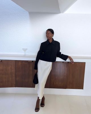 Sylvie wears a short black jacket, white slip skirt, black heels, and a black bag.