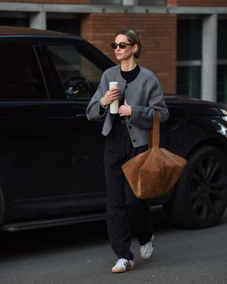 Anouk wears a gray short jacket, a black top, black pants, white Adidas Sambas, and a brown suede tote bag.