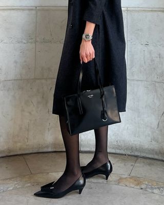 Eliza Huber wearing a gray coat, silver IWC watch, tights, Prada kitten heel pumps, and a 1995 Prada rectangular bag.