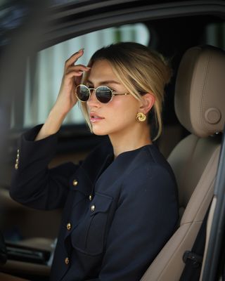 female fashion influencer and accessory designer Cass DiMicco poses in a car with Aureum wire-rim oval sunglasses, statement yellow gold earrings, and a navy lady jacket with gold buttons