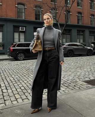 female fashion influencer and accessory designer Cass DiMicco poses on a NYC sidewalk wearing a long gray coat, gray ribbed turtleneck, tan bag, Aureum belt, wide-leg black jeans, and brown pumps