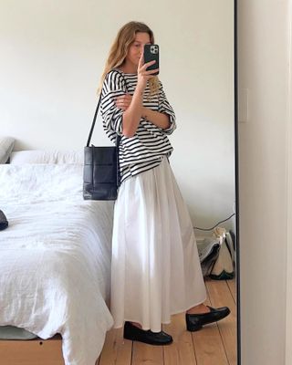 British female fashion influencer Brittany Bathgate poses for a mirror selfie wearing an oversize striped long-sleeve t-shirt, woven black Bottega Veneta bucket bag, white full skirt, and black loafers