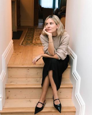 British female fashion influencer Lucy Williams poses sitting on the stairs in her London home wearing a neutral sweater, black midi skirt, and pointed-toe black slingback flats