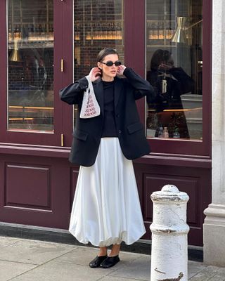 female fashion influencer Valeria sobalera poses on a London sidewalk wearing black oval sunglasses, an oversize black blazer, black sweater, white bubble-hem full skirt, and black glove flats