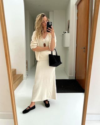 female fashion influencer Anouk Yve poses for a mirror selfie wearing a long pendant necklace, cream v-neck sweater, knit maxi skirt, black mini tote bag, and black suede Alaia Mary Jane flats