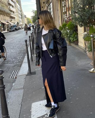 French fashion influencer Anne-Laure Mais poses on a Paris sidewalk wearing black sunglasses, a black leather jacket, white t-shirt, navy pinstripe maxi skirt, white crew socks, and black Adidas Samba sneakers