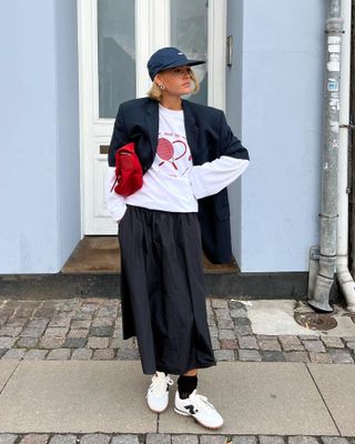 Scandi Danish female fashion influencer Amalie Neilsen poses on a sidewalk in Copenhagen wearing a navy baseball cap, oversize navy blazer, longe-sleeve tennis graphic tee, black full skirt, red cluthc, black scrunched ankle socks, and white New Balance sneakers