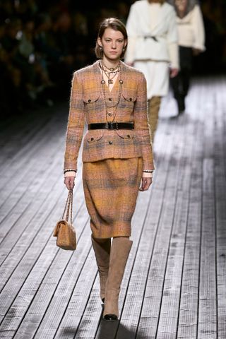 A Chanel F/W 24 look including a pink-and-yellow plaid collarless blazer with a black belt and matching pencil skirt with cap-toe knee-high boots.