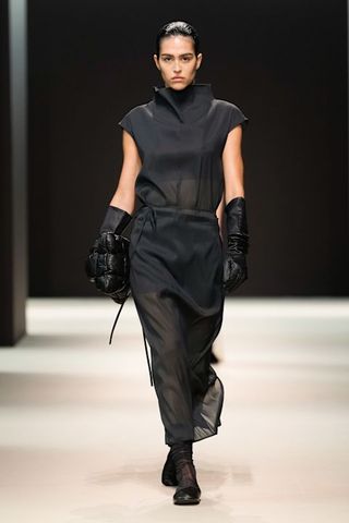 A woman at COS F/W 24 show wearing a sheer blouse, sheer skirt, leather gloves, a padded clutch, and sheer socks with ballet flats.