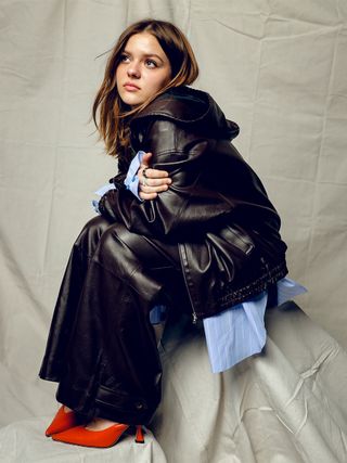 Maisy Stella wears leather jacket and pants with oversize blue button down shirt and red heels.