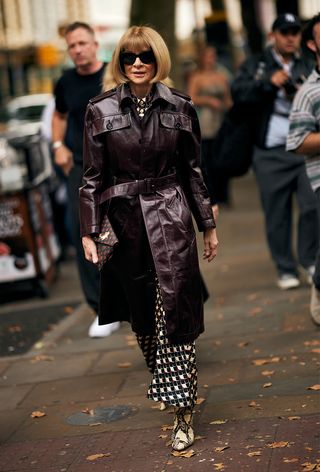 a street style photo of Anna Wintour wearing a brown leather trench over a printed dress with boots to the JW Anderson spring 2025 runway show