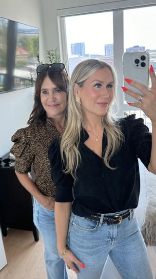 A woman wearing a black blouse with jeans and a statement belt taking a selfie with a woman wearing a leopard print blouse.