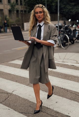 A woman wearing geek chic trend in the form of a gray blazer and matching skirt with a white button-down shirt, a black tie, and black pumps during Milan Fashion Week spring/summer 2025.