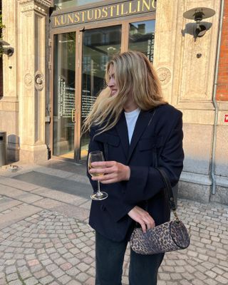 Danish female fashion influencer Sidsel Alling poses on a sidewalk in Copenhagen, Denmark holding a glass of wine wearing a black blazer, white tee, a leopard-print mini duffel bag, and black jeans