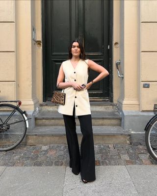 British female fashion influencer Anna Newton poses on a London sidewalk wearing a mini leopard-print bag, long cream vest top, and black flared pants