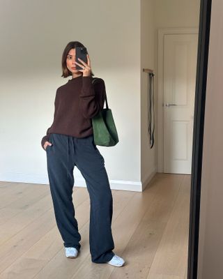 A woman wears a chocolate brown sweater, navy pants, white sneakers, and a green suede tote bag.