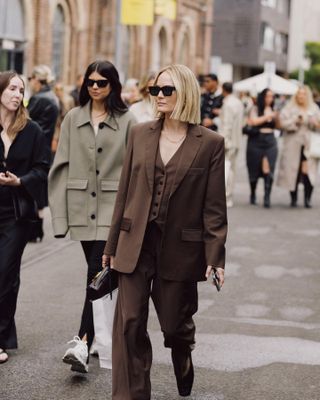 A woman wears a chocolate brown suit, vest, pants, and black shoes with black sunglasses.