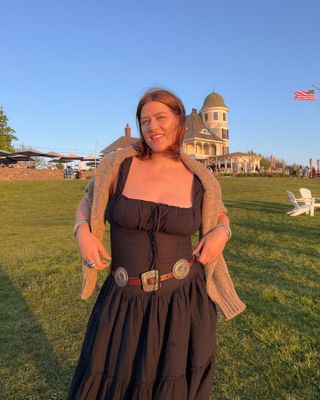 Rae wears a chocolate brown dress with a tan sweater, and a western-inspired belt.