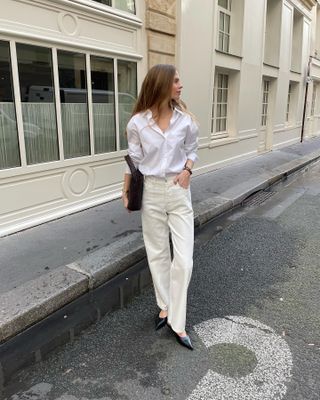 Clara wears a white button-down shirt, ivory jeans, black flats, and a black bag.